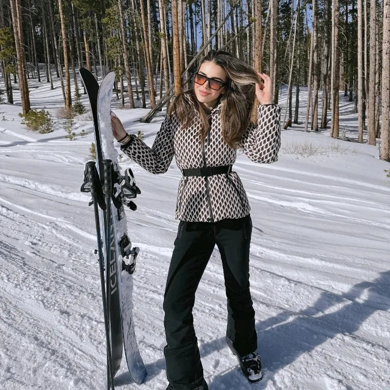A woman in a geometric print, belted ski jacket and black trousers stands in the snow holding a pair of skis.