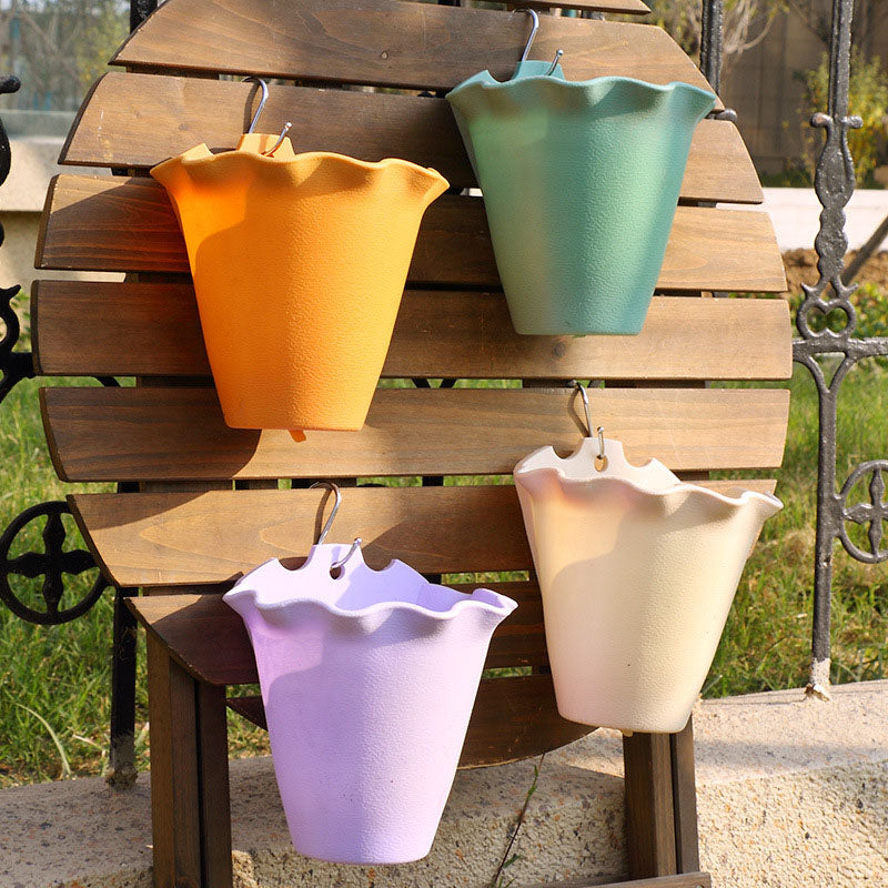Four pastel-colored, scalloped-rim plastic planters hang from silver hooks on a wooden slatted outdoor wall display.
