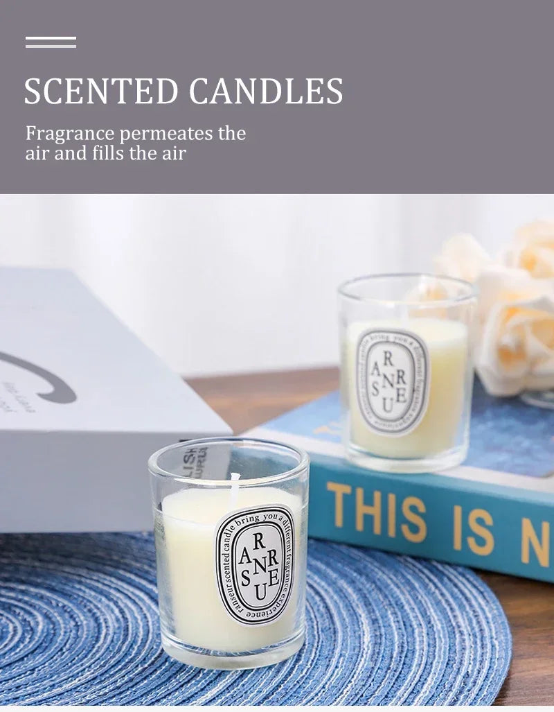 Two small, cream-coloured scented candles in clear glass tumblers with oval labels, sitting on a blue woven placemat.