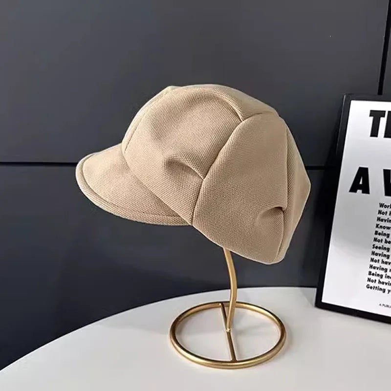 Beige retro newsboy beret hat with a structured peak, displayed on a gold stand against a dark grey background.