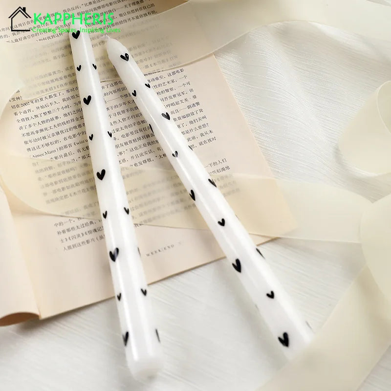 Two white tapered dinner candles decorated with small, black heart patterns, resting on paper and ribbon.