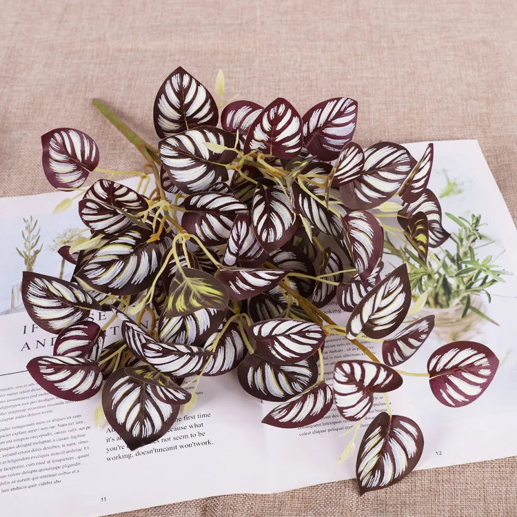 Artificial plant branch with dark purple oval leaves featuring striking white striped patterns on a white paper background.