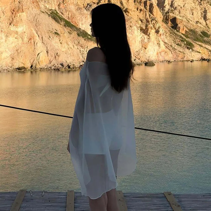 Woman wearing a sheer, white, off-the-shoulder beach cover-up, standing on a wooden pier by the sea.