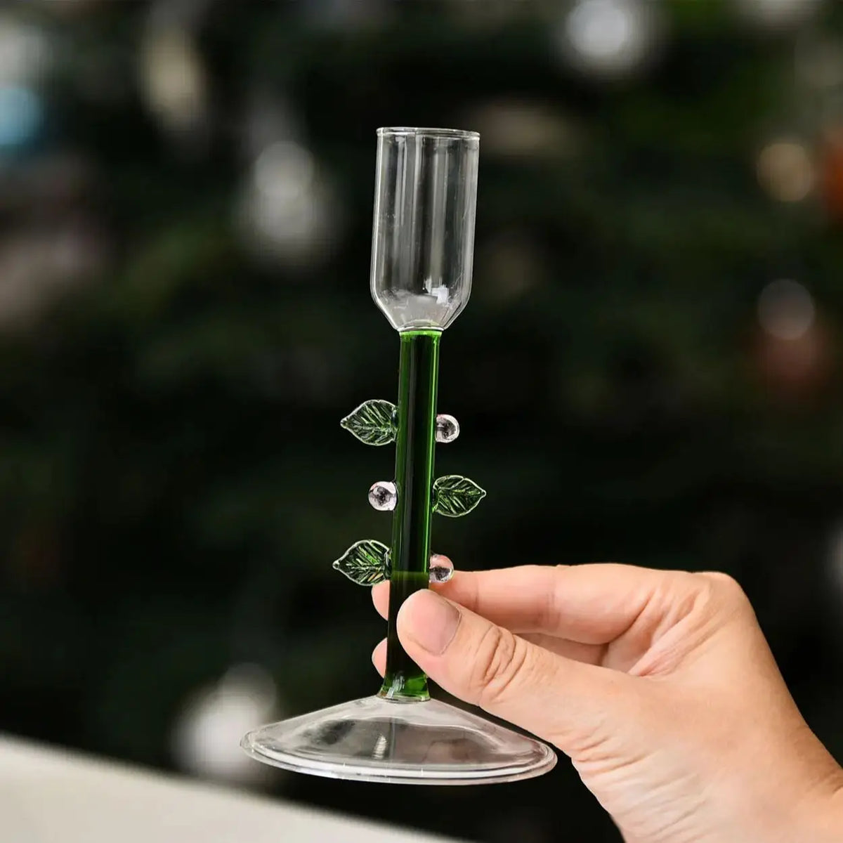 Clear borosilicate glass candlestick holder with a green stem, decorative leaves, and berries held by a hand.