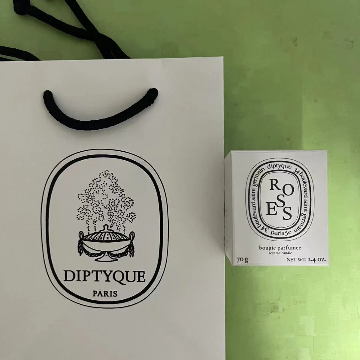 White gift bag with black logo next to a 70g "Roses" scented candle box. Minimalist white and black design.