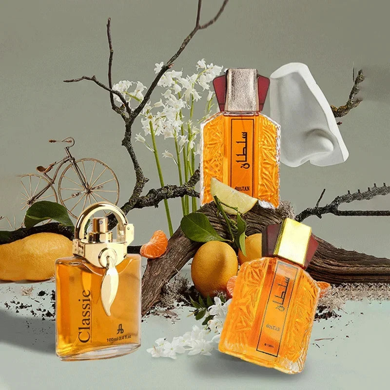 Three amber perfume bottles among twigs, citrus fruit, and white flowers, with a small bicycle and nose sculpture.