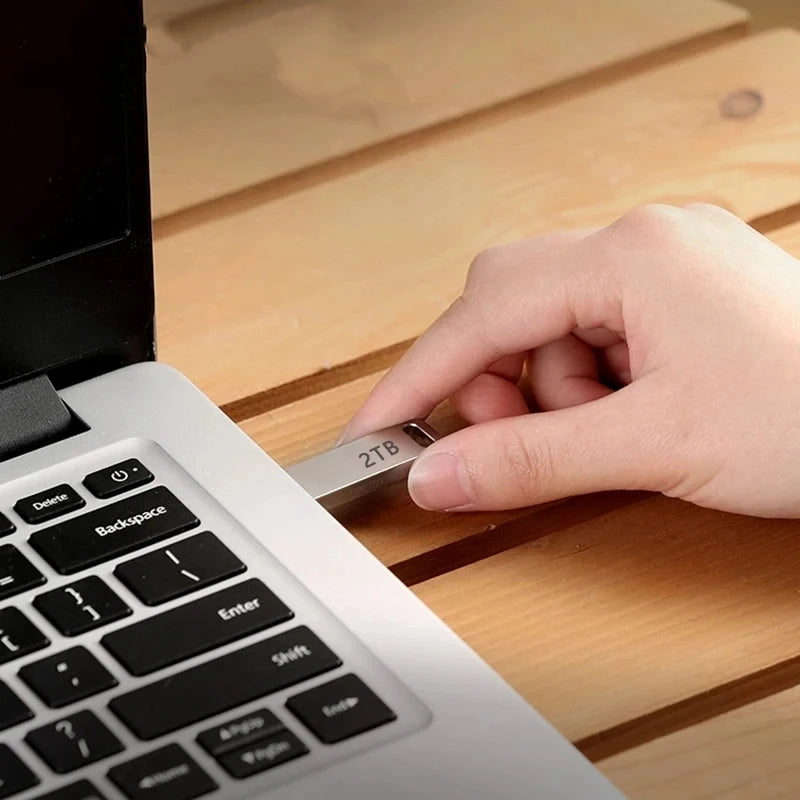 Hand inserting a sleek, silver 2TB metal USB flash drive into a laptop port on a wooden desk.