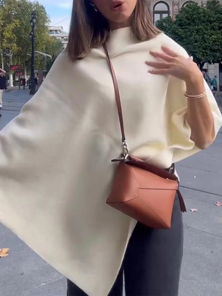 Cream-colored draped knit poncho with an asymmetrical hem and high mock neck, worn with a brown crossbody bag.