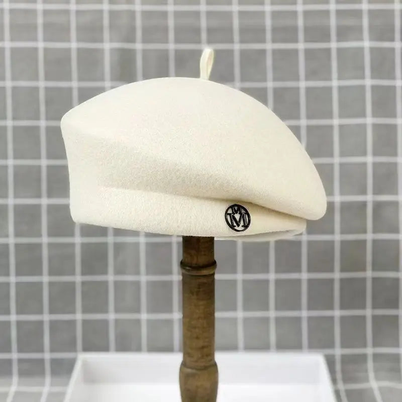 Classic cream-coloured wool felt beret with a small, circular metallic logo detail on the side.