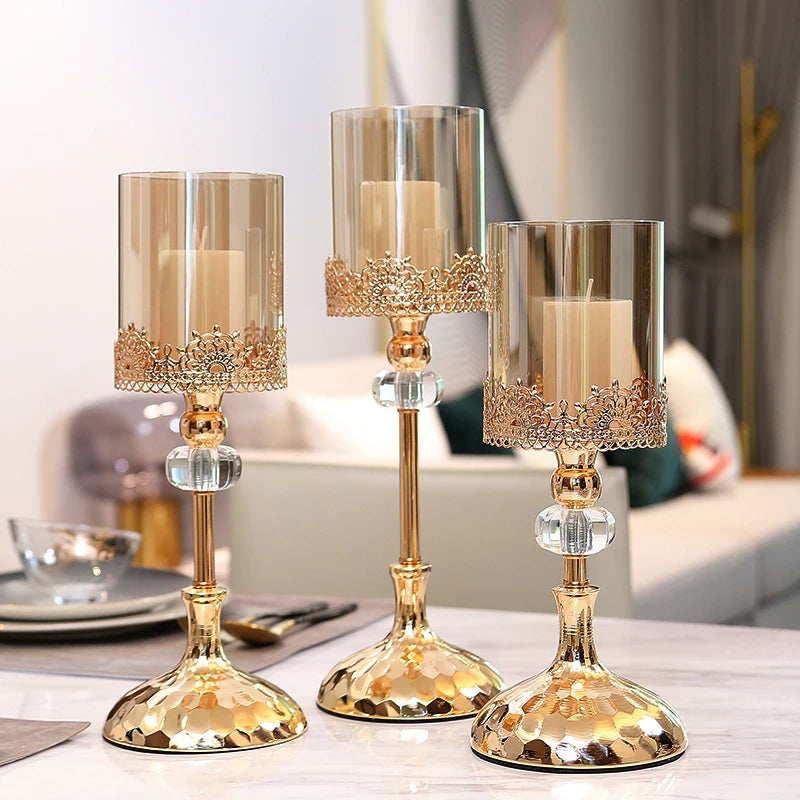 Three gold crystal candle holders with glass cylinders and pillar candles on a table.