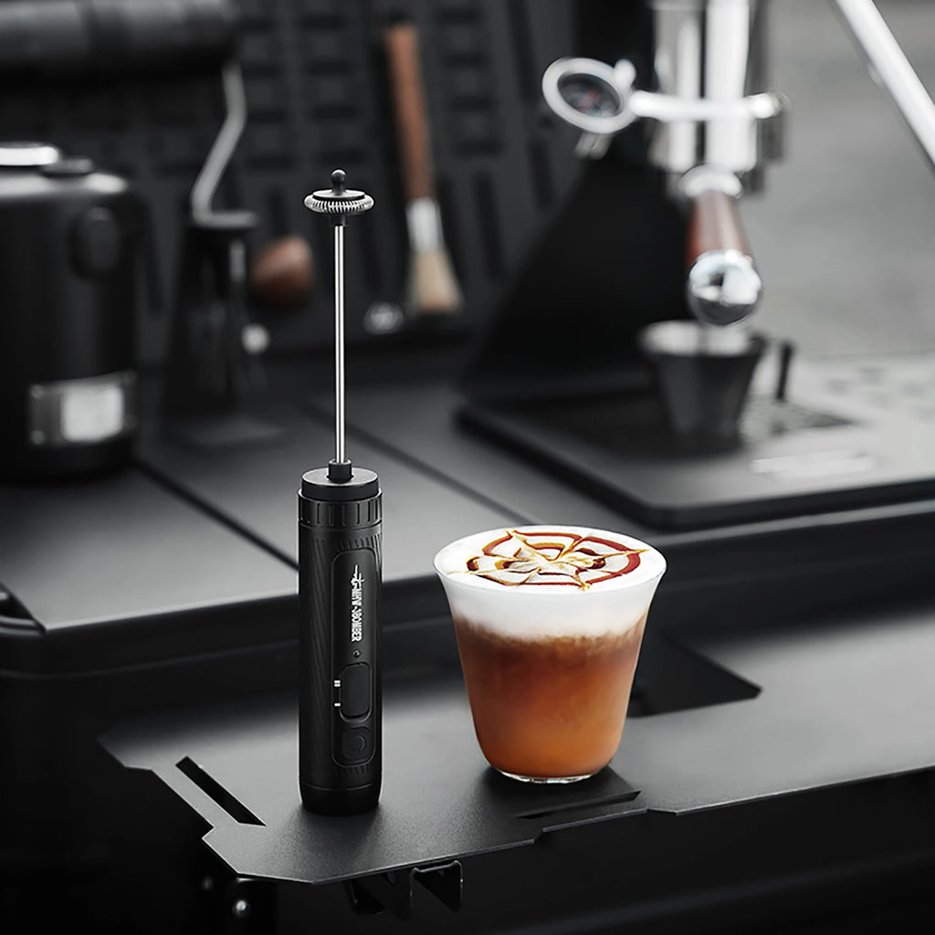 A sleek, black handheld electric milk frother with a long metal whisk, standing next to a glass of latte art coffee.
