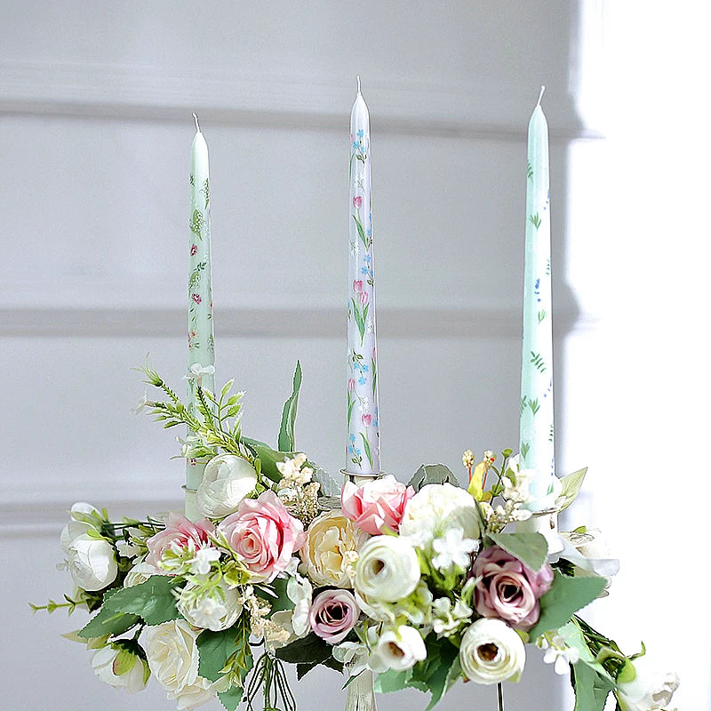 Three pastel taper candles featuring floral prints, arranged elegantly in a lush bouquet of white and pink faux roses.