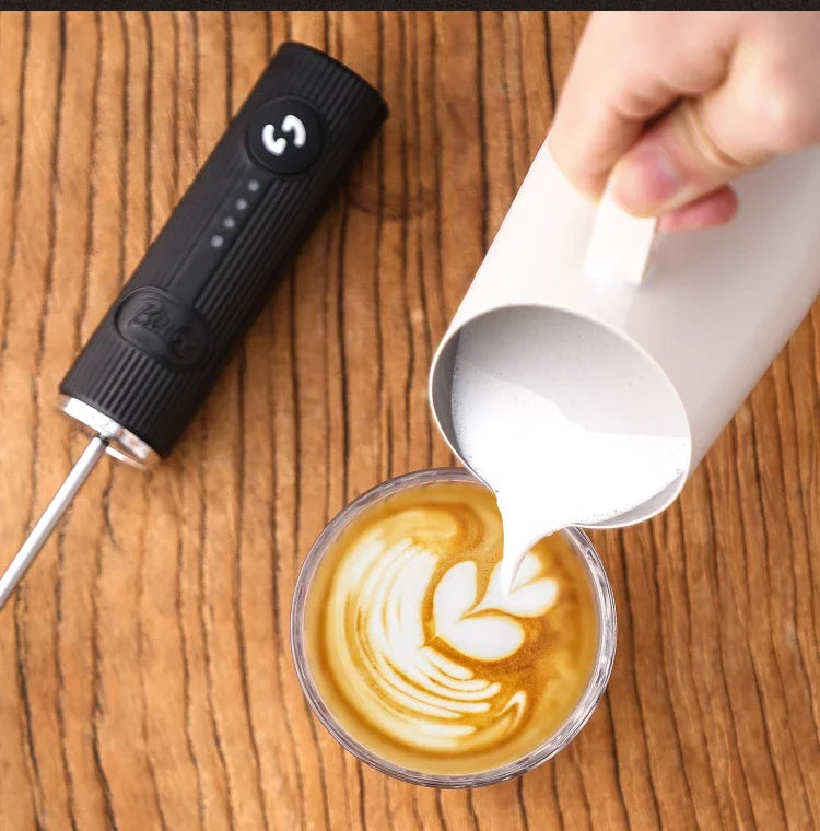 Handheld black electric milk frother next to a latte with heart-shaped foam art being poured from a white pitcher.