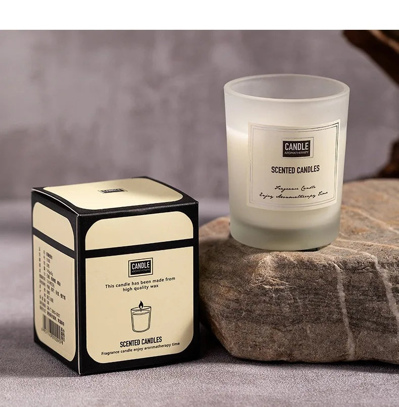 A white scented candle in a frosted glass holder with its matching cream and black gift box on a stone surface.