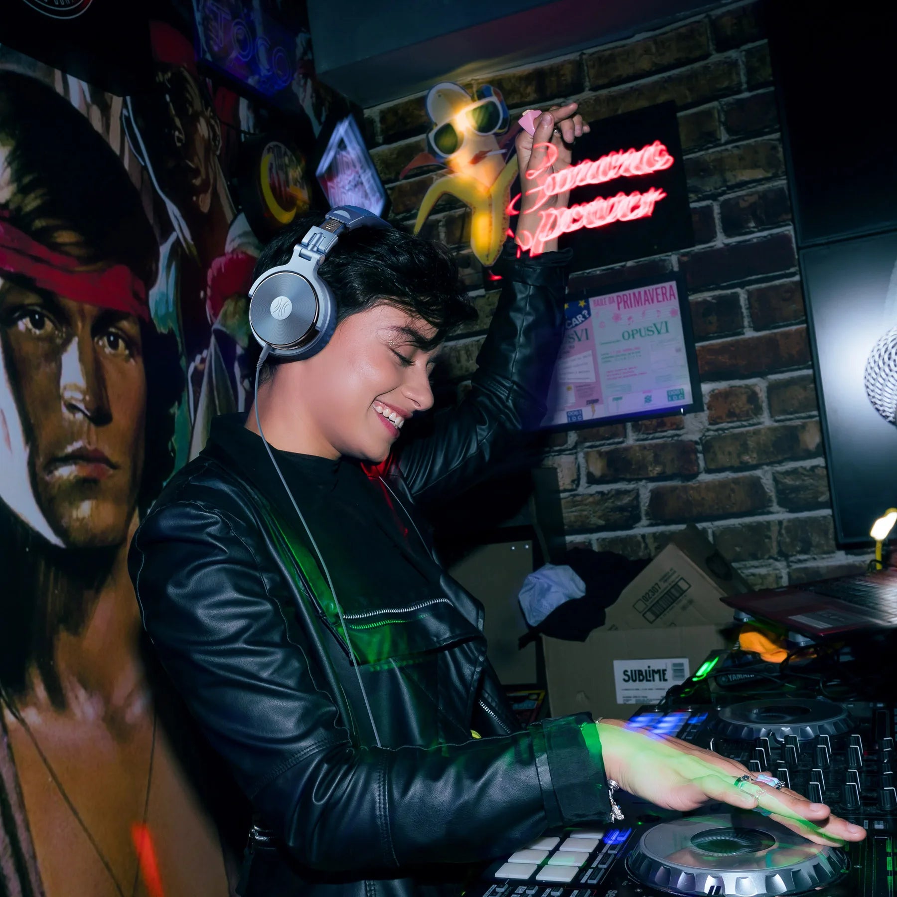 Professional silver wired DJ studio headphones with an integrated microphone, worn by a smiling DJ at a console.