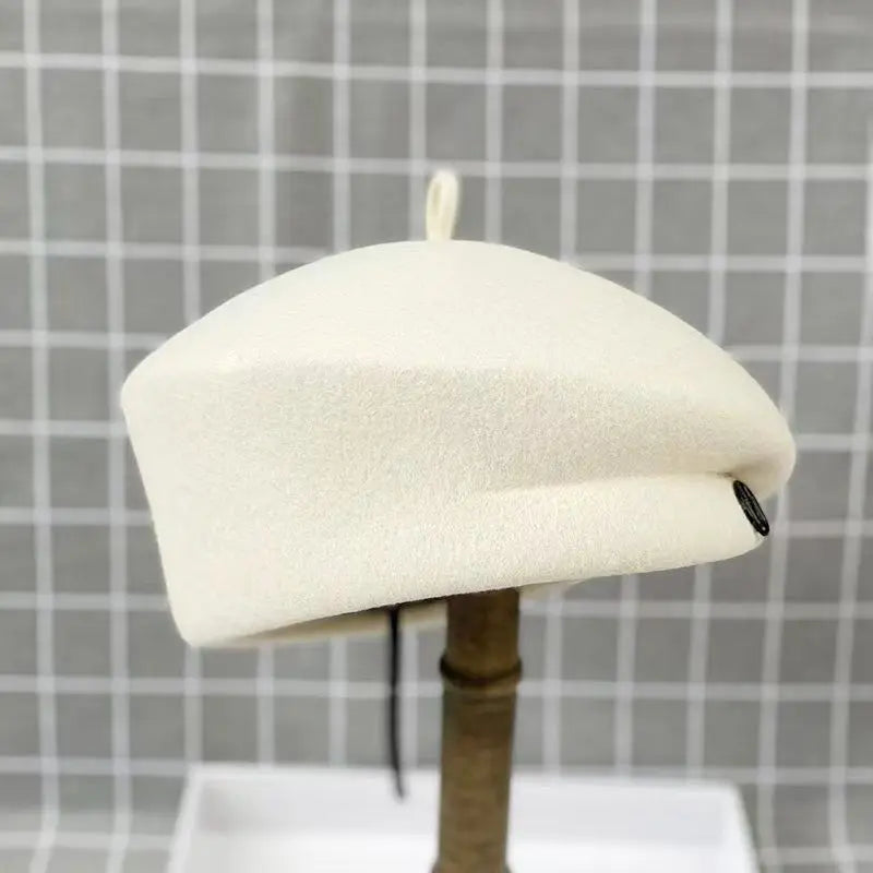 Cream-coloured wool felt beret with a small top loop and side detail, displayed on a stand against a grid background.