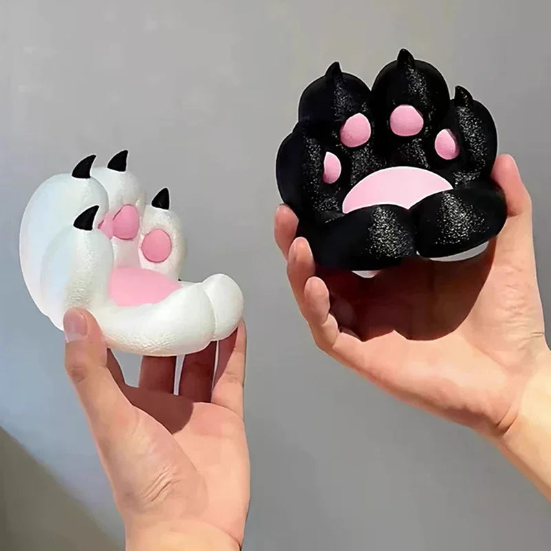 Two hands holding a white and a black cat paw squeeze toy with pink pads and contrasting claw details.