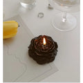 A dark brown, rose-shaped candle with a glowing flame, sitting on a table next to a yellow flower.
