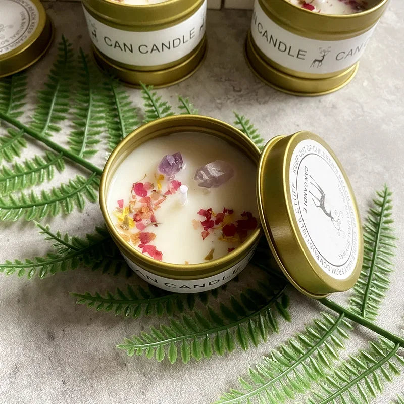 Scented soy candle in a gold tin, topped with dried flower petals and small amethyst crystals on a fern leaf background.