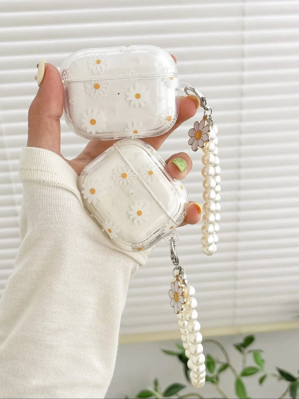 Clear daisy-print AirPod case with faux pearl wrist strap and flower charm, held in a hand.