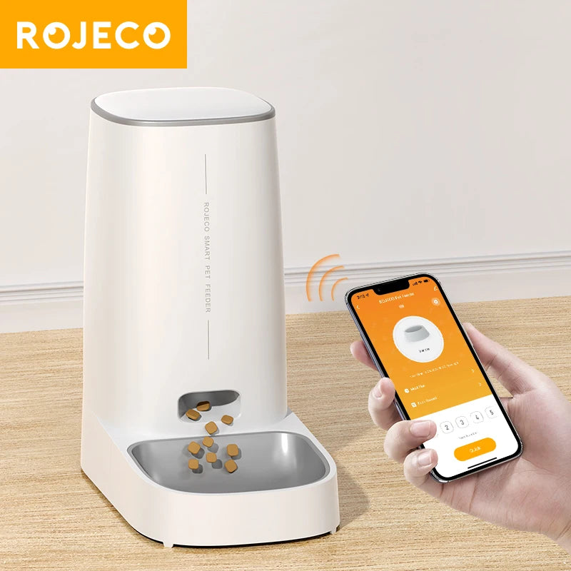 White smart pet feeder with a metal bowl, controlled by a smartphone app shown in a hand. Modern, minimalist design.