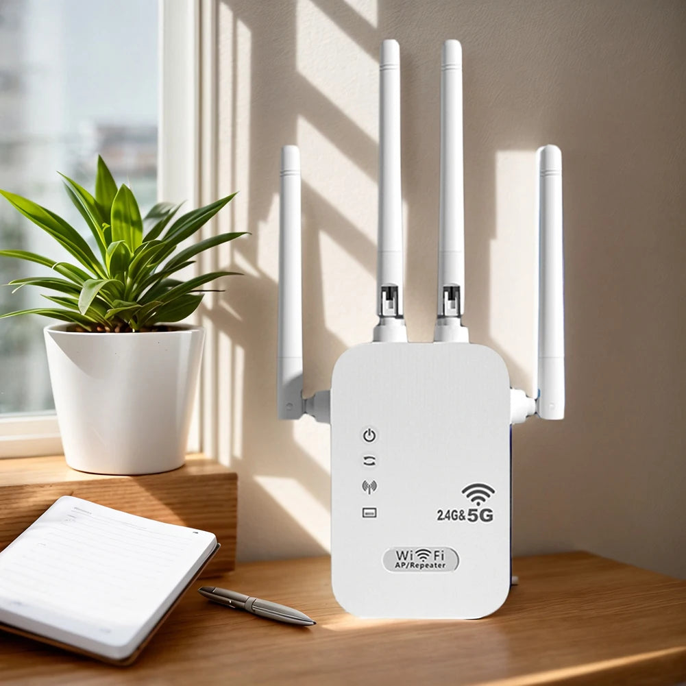 White dual-band WiFi repeater with four external antennas, sitting on a wooden desk next to a plant and notebook.