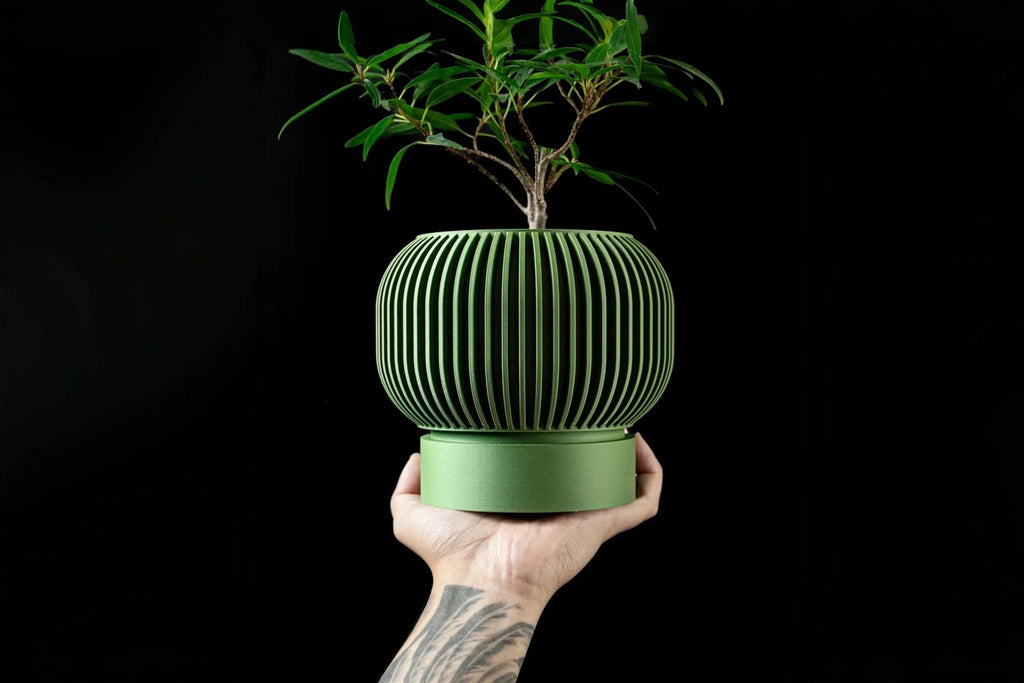 A hand holds a minimalist, olive-green 3D-printed plant pot with vertical slats, containing a small leafy plant.