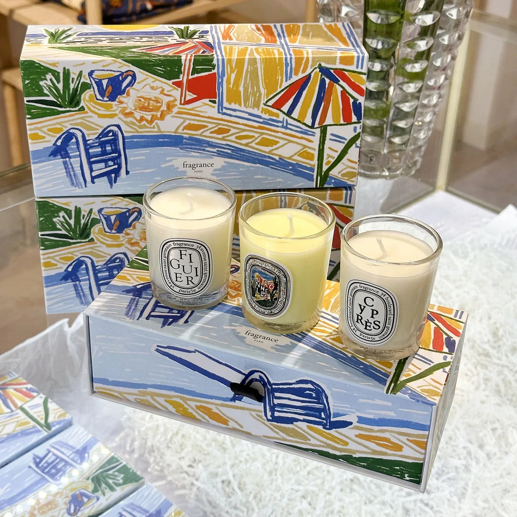 Three scented candles in glass jars displayed on a colourful, summer-themed illustrated gift box.