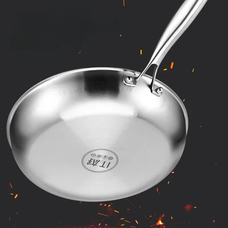 A stainless steel frying pan with riveted handle and branded bottom, angled against a dark background with orange sparks.