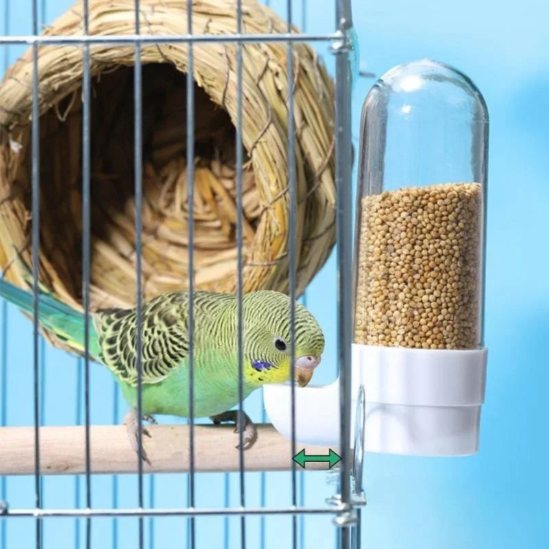 A parakeet eats from a clear, white plastic bird feeder attached to a cage near a woven straw nesting hut.