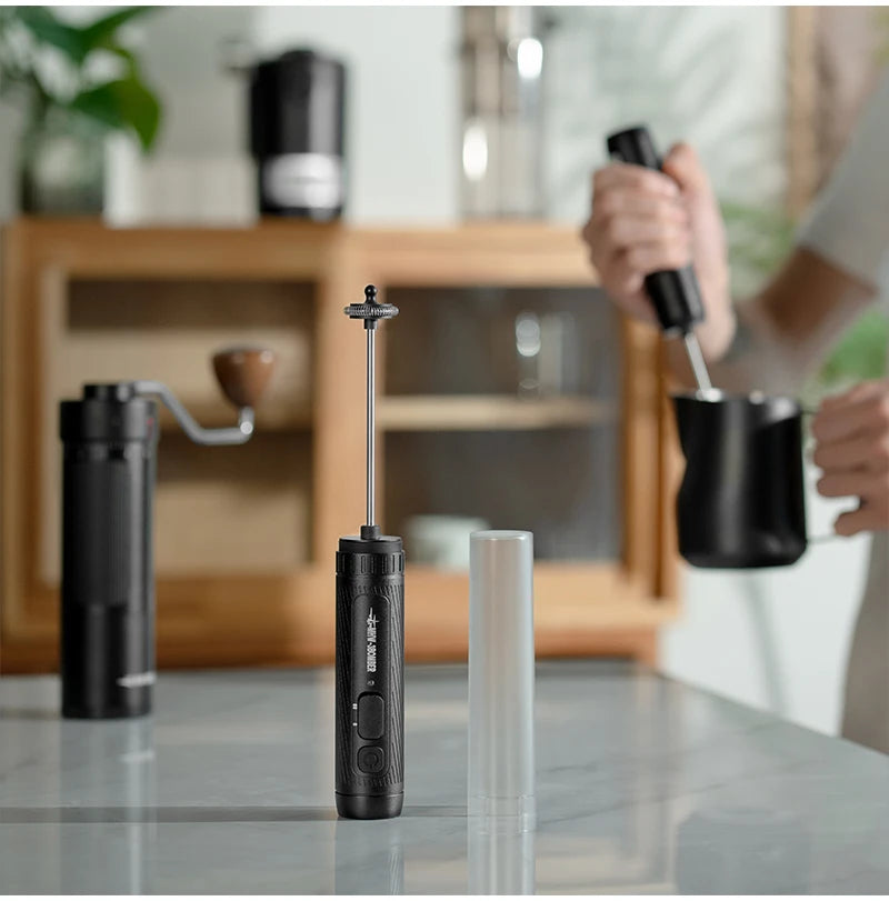 Black handheld electric milk frother with a slim metallic whisk, presented next to its clear protective cover.