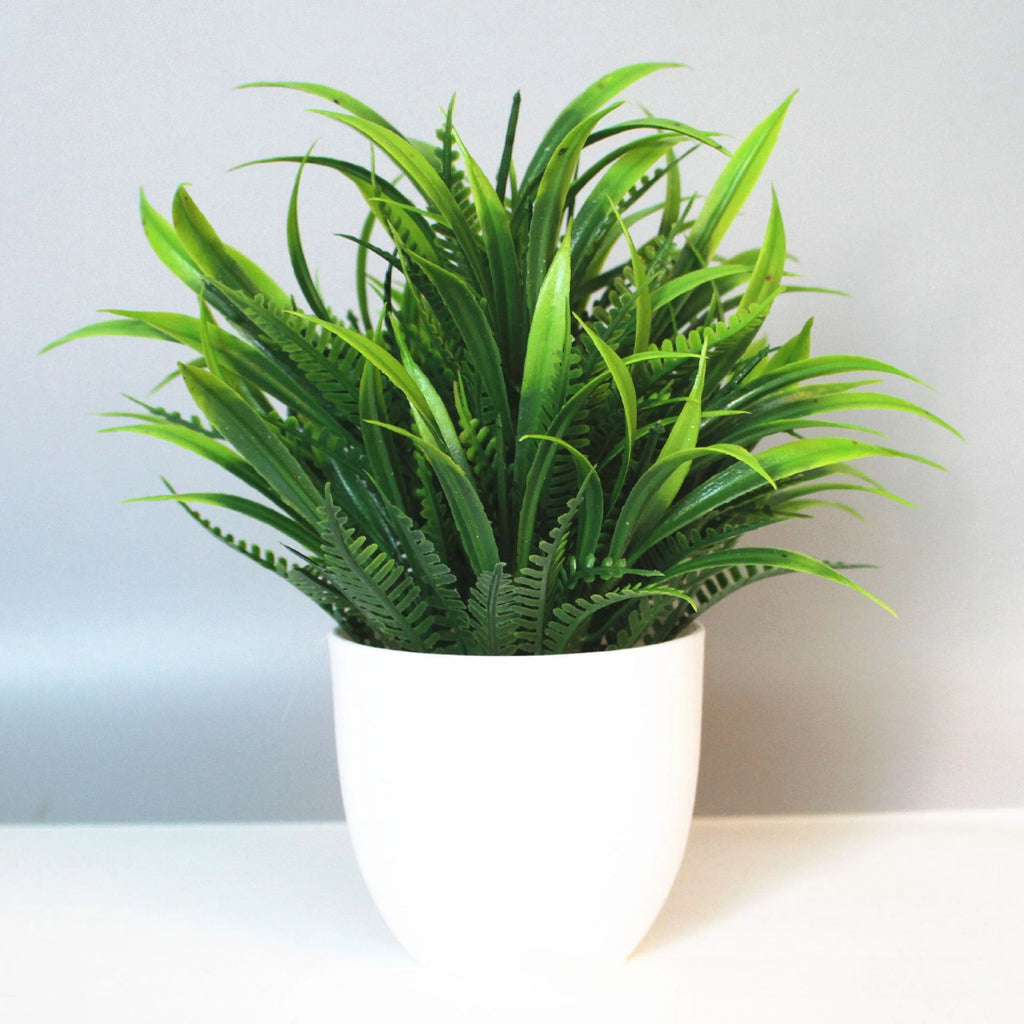 Artificial plant with spiky green leaves in a simple, minimalist white rounded pot against a light gray background.