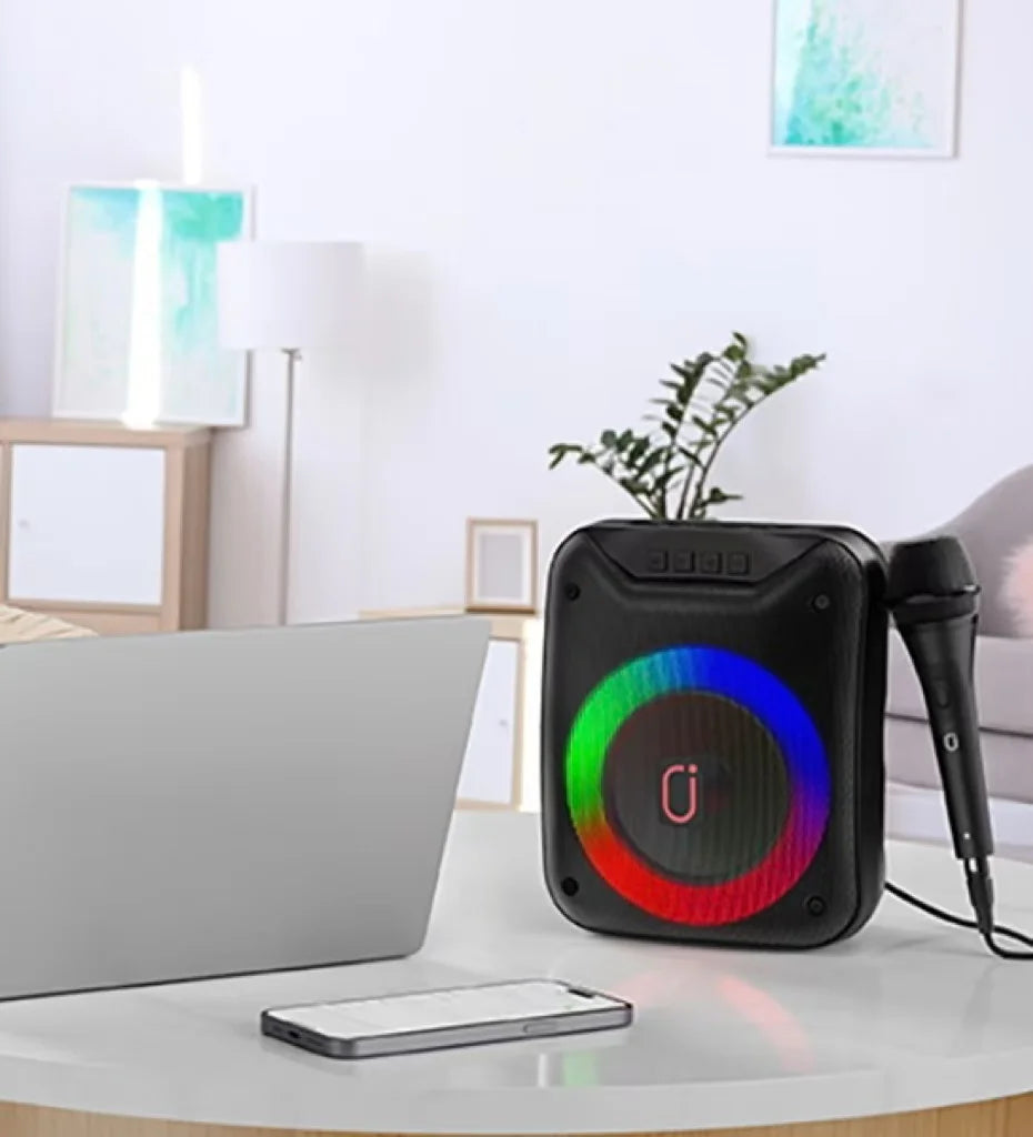 Portable black wireless speaker with a circular multi-coloured LED light and a wired microphone on a table.