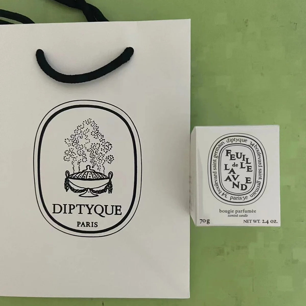 A white 70g scented candle in its box next to a matching branded white gift bag with black handles.