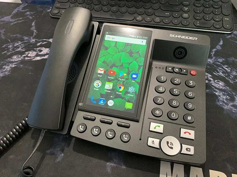 Black IP desk phone with a large touchscreen display, numeric keypad, handset, and integrated video camera.