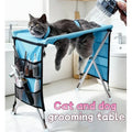 Foldable blue pet grooming table and hammock with metal legs, mesh side pockets, and grooming tools.