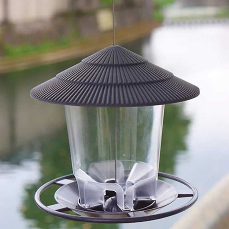Hanging bird feeder with a dark brown, ridged roof, clear cylindrical seed container, and a circular feeding perch.