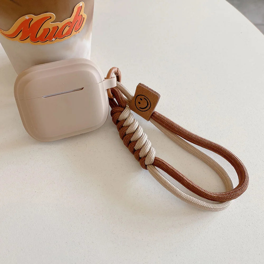 Beige silicone AirPods case with a braided two-tone brown and cream wrist strap featuring a smiley face tag.