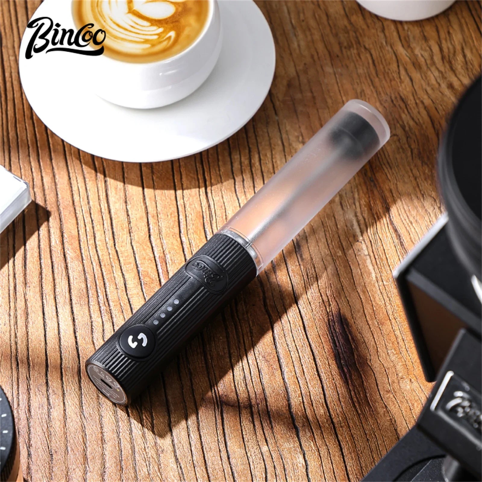 Portable black electric milk frother with a translucent upper shaft, placed on a rustic wooden table.