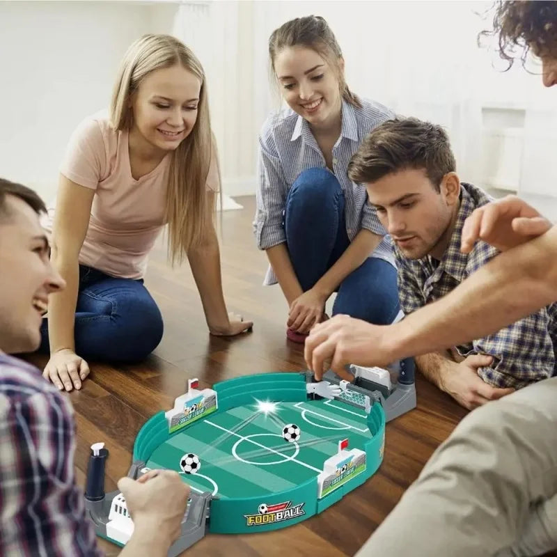 Group of friends playing a green tabletop football game with two balls and manual control handles on a wooden floor.