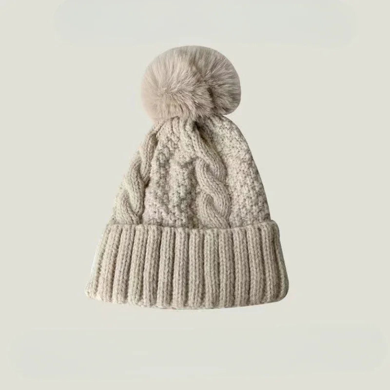 Beige cable-knit winter beanie featuring a ribbed cuff and a fluffy faux-fur pom pom on top.