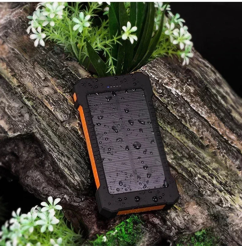 Rugged black and orange portable solar power bank, wet from rain, resting on a wooden log with white flowers.