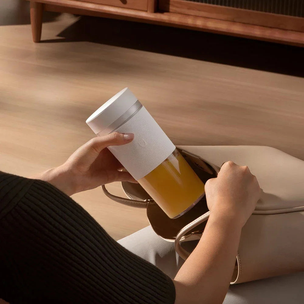 A person places a white, ribbed portable blender with yellow juice into a beige handbag.
