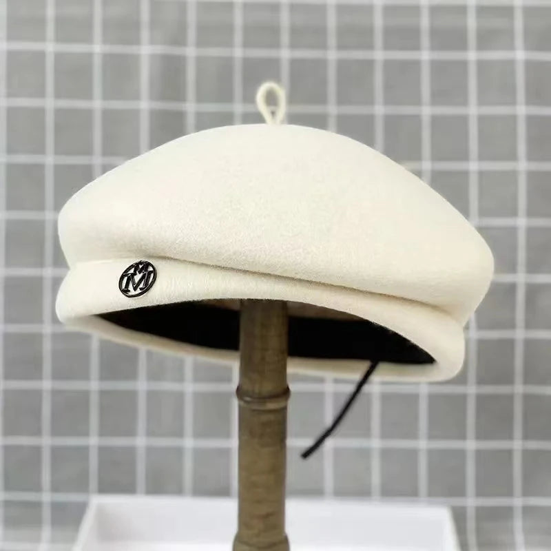 Classic cream wool-felt beret with a small top loop and a decorative circular metal emblem on the side.