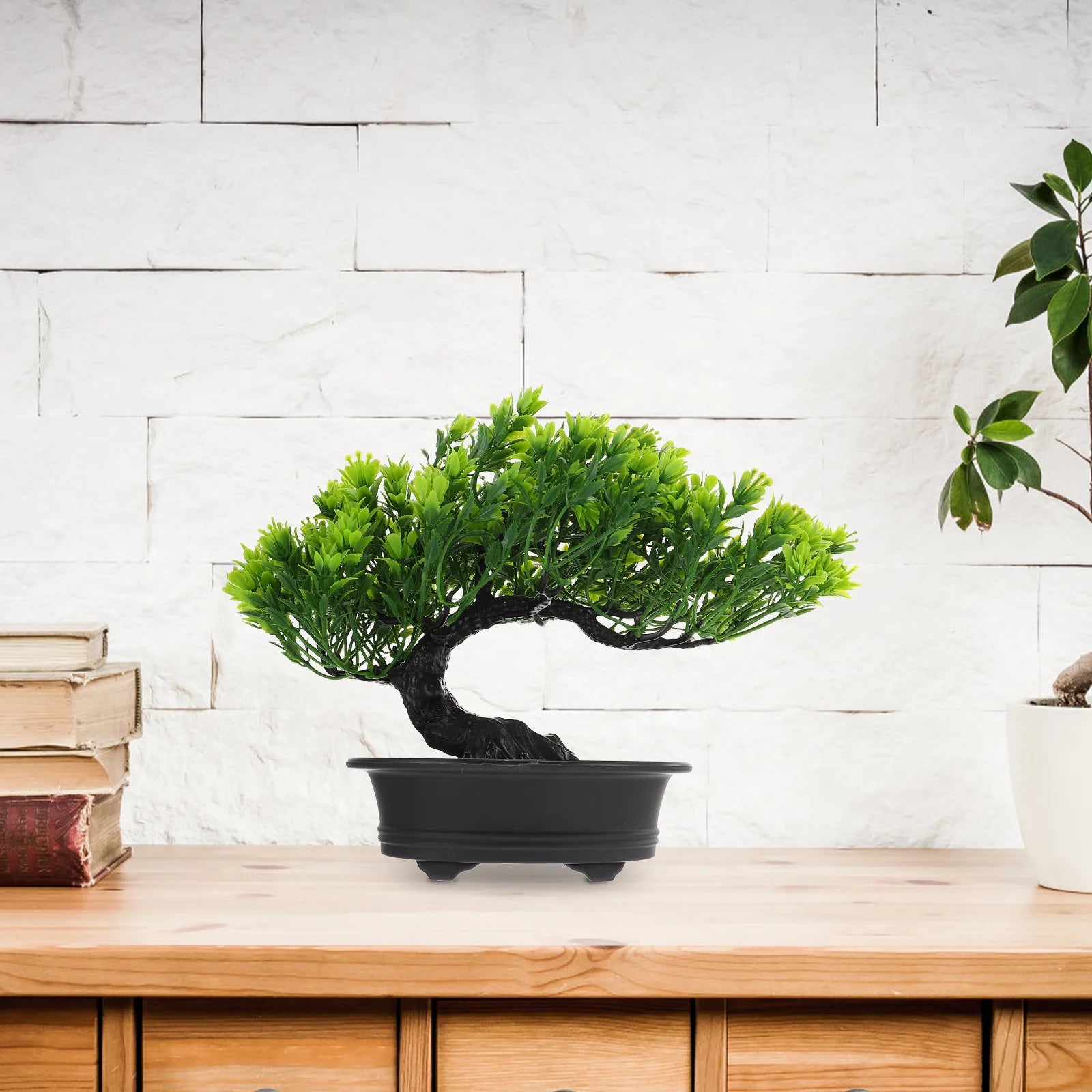 Artificial green bonsai tree with a curved black trunk in a rectangular black pot, sitting on a wooden table.