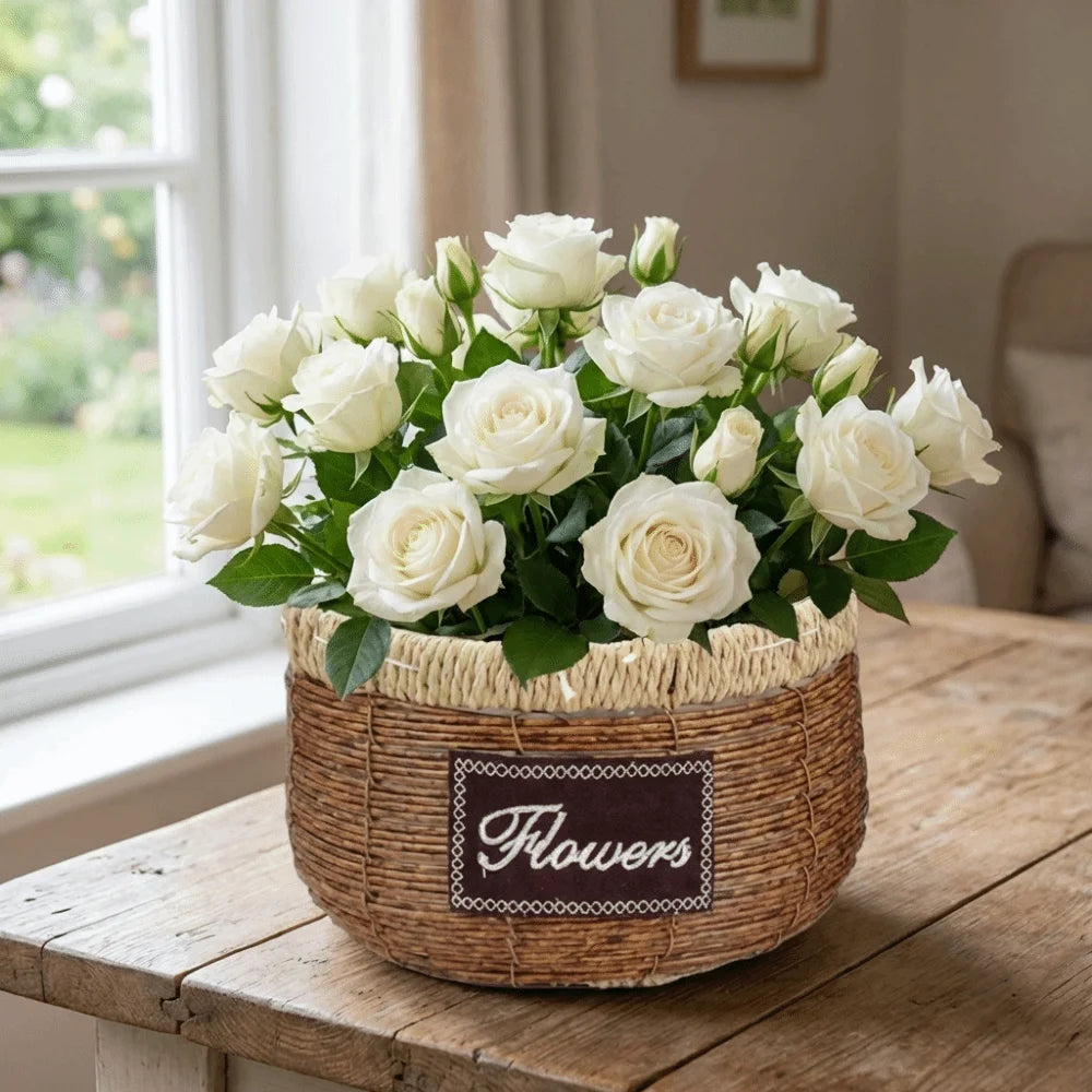 A bouquet of white roses in a rustic brown woven basket with a "Flowers" label, sitting on a wooden table.