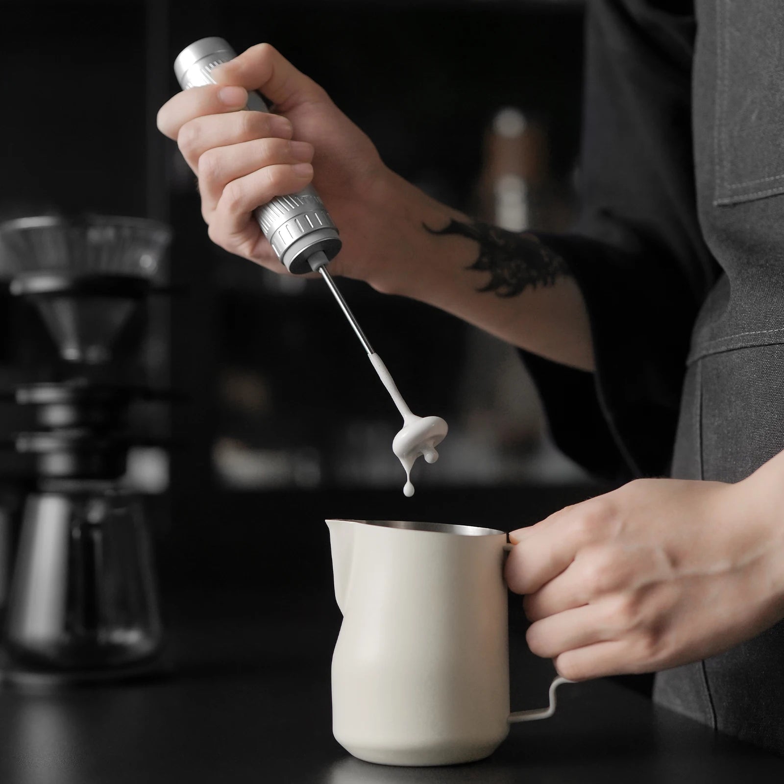 A person uses a handheld silver electric milk frother over a small, matte cream-colored metal pitcher.