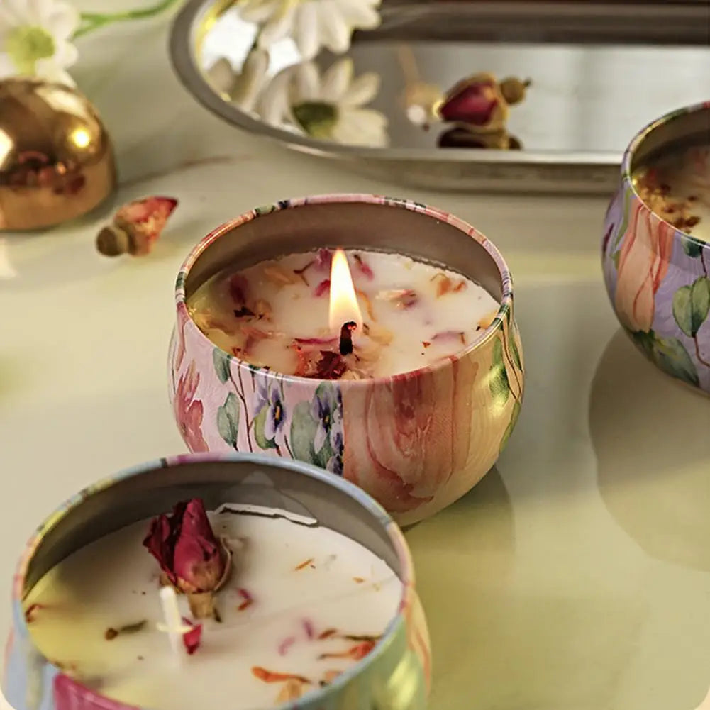 Lit scented candles in colourful, floral-patterned metal tins, decorated with dried flower petals.