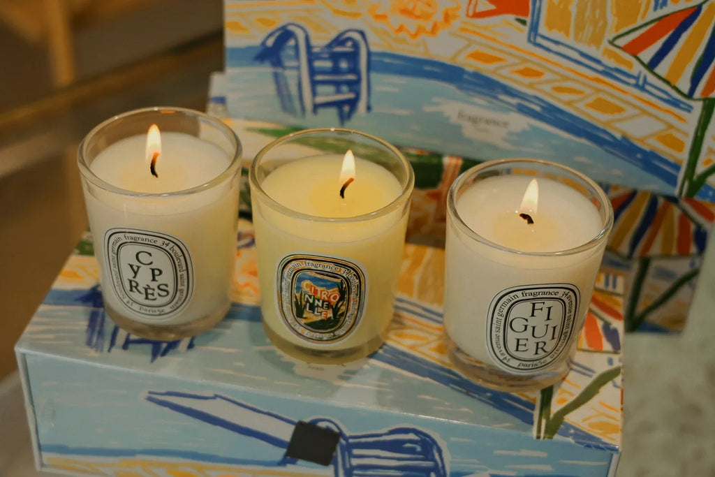 Three lit scented candles in glass jars with white labels, presented on a decorative illustrated gift box.
