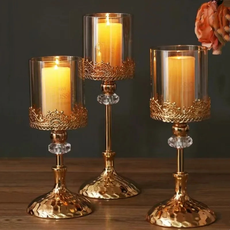 Three gold and crystal candle holders with glass cylinders and lit pillar candles on a wooden table.